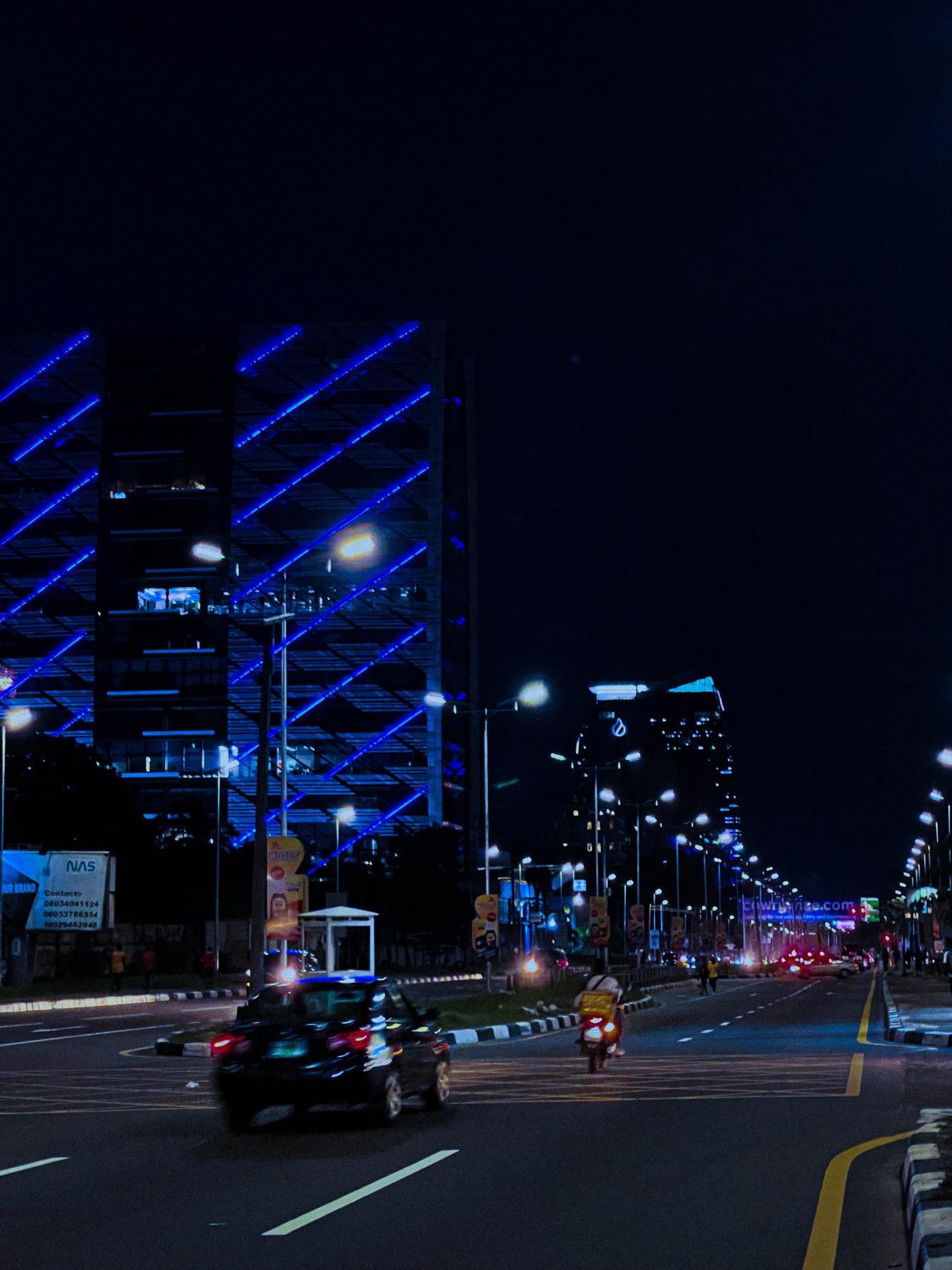 A rider passing a blue-lit building on a Lagos road at night